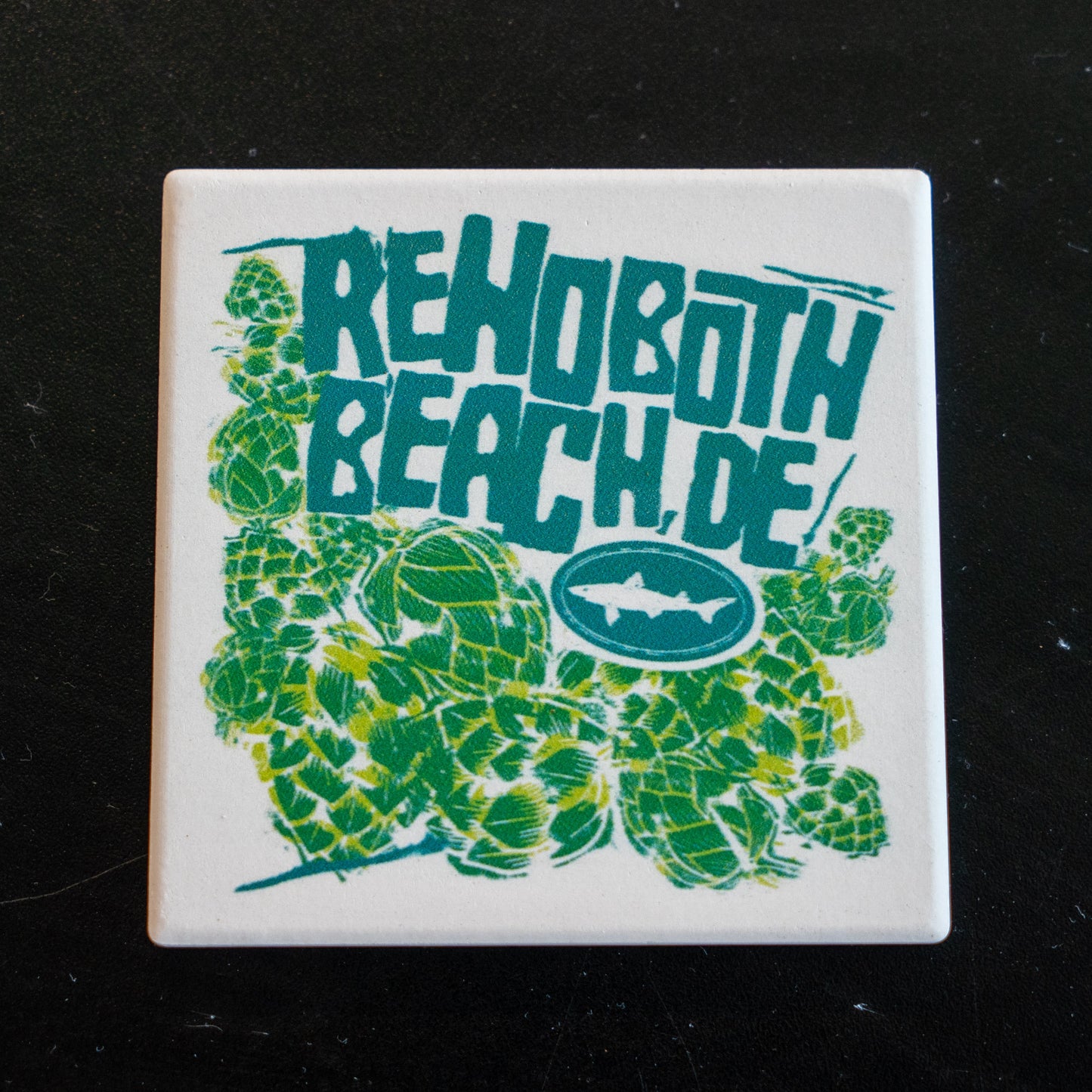 The B&E Coaster by Paint the Town, Inc. is a square stone coaster with green hop designs, a small fish logo, and bold teal "Rehoboth Beach, DE" text on white—made from absorbent coaster stone and ideal for any black tabletop.