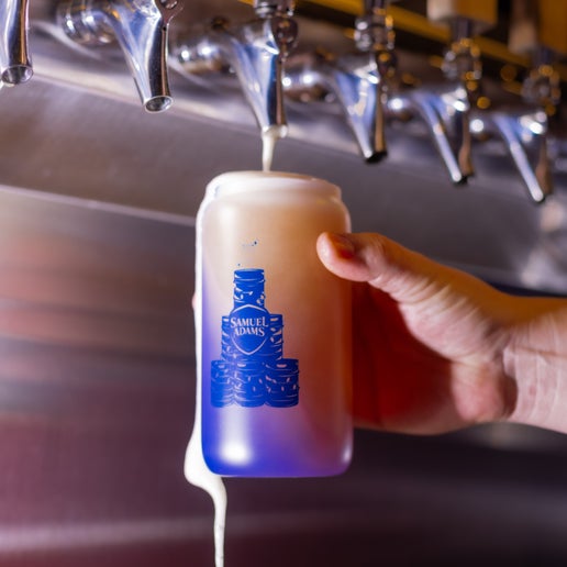 A frosted glass with a pyramid of kegs wrapped in Christmas lights with a perfect pint glass sitting on top and the Samuel Adams classic shield logo in the middle in navy printed on the other side of it that changes color to a blue when it comes into contact with cold. In this image the glass has just been filled with beer from a tap and is actively changing color to blue.