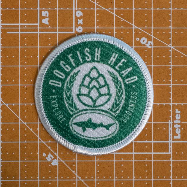 A round green Explore Goodness Patch by GRAFFITI INC features white embroidery with "Dogfish Head" and "explore goodness," plus a hop cone, laurel branches, and fish logo—perfect for inspiring adventure in every sip.
