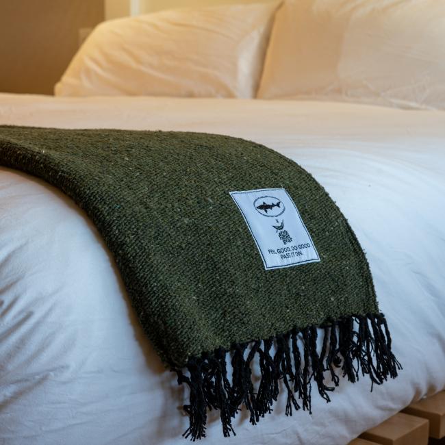 The Trek Light Gear DFH INN Blanket, dark green with a white patch, is draped over the edge of a bed made with white bedding in a softly lit room.