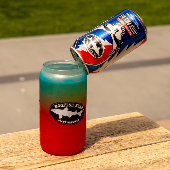 A vibrant Ombre Color Changing Can Glass from Grandstand Glassware & Apparel sits on wood, topped with a can of blueberry vodka soda. The background is grassy and blurred, making the colorful drink stand out.