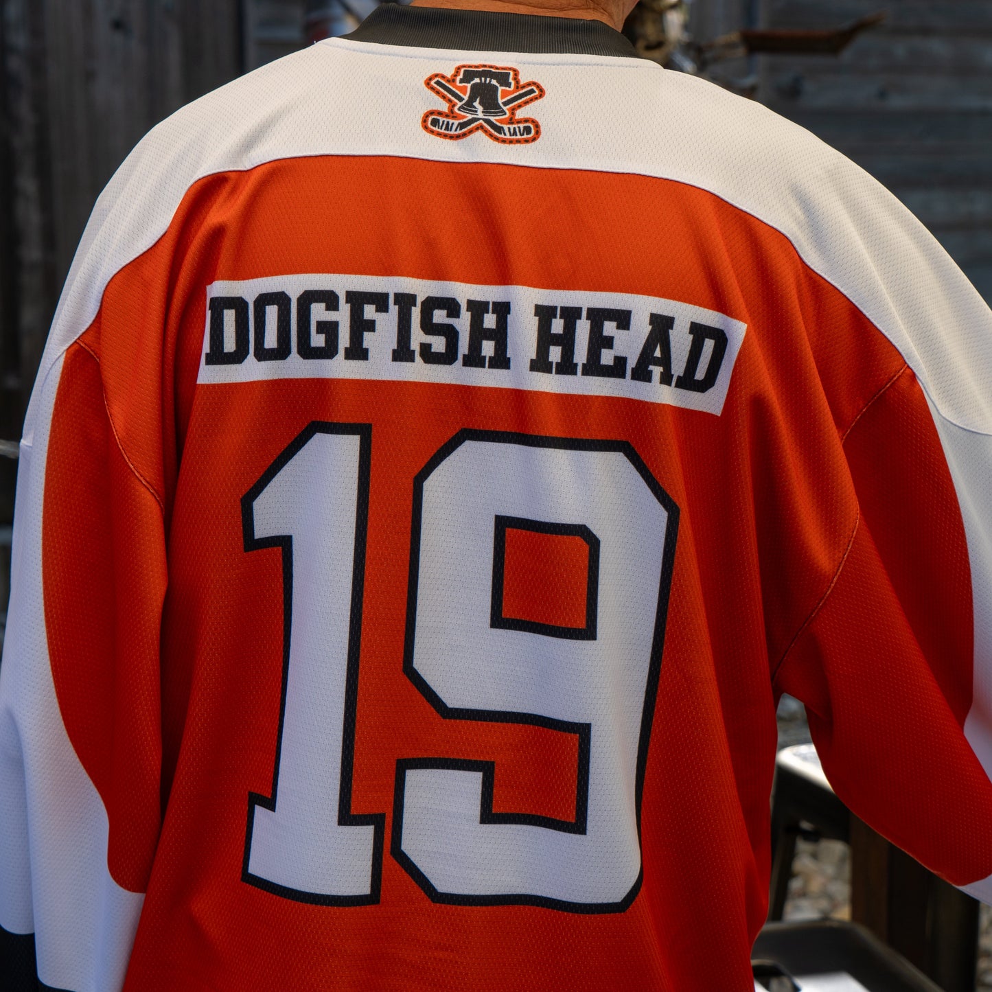 Someone wearing an orange, white, and black Dogfish Head x Garnet Hathaway Hockey Jersey by In Your Face Apparel, featuring the number 19, DOGFISH HEAD on the back, and a logo by the collar.