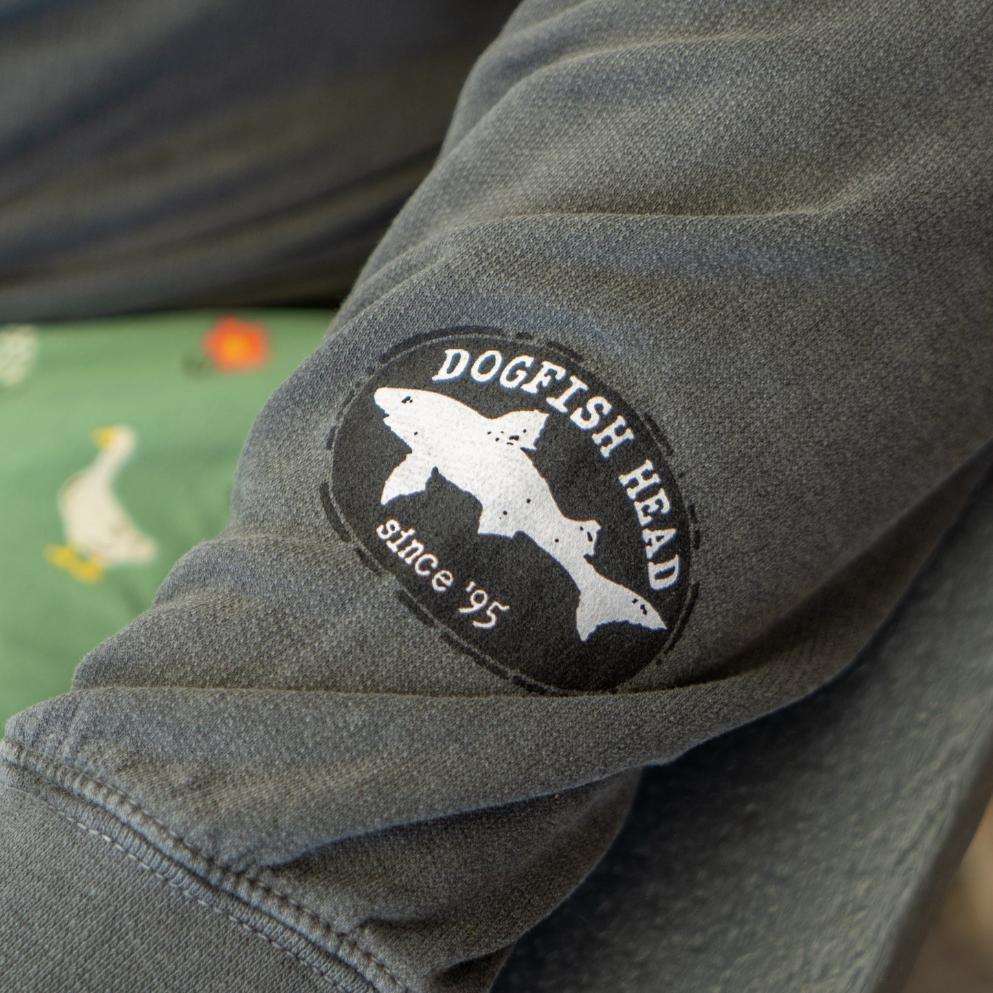 A close-up of the Grey Got A Minute Hoodie by GO WEST T-SHIRT COMPANY shows its sleeve with a Dogfish Head since 95 patch. In the background, green fabric with duck or goose illustrations appears. Perfect for beer apparel fans.