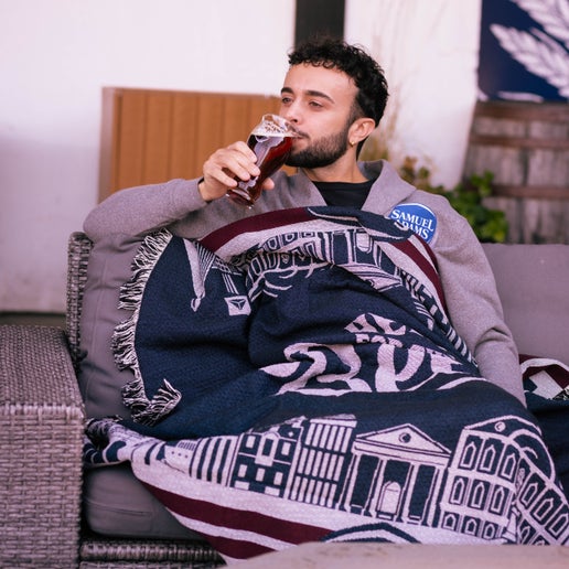 A 72" x 55" blanket that features the Boston skyline in white over a navy background and features the phrase "Here for the beer" in the center in white letters. Pictured here in use.