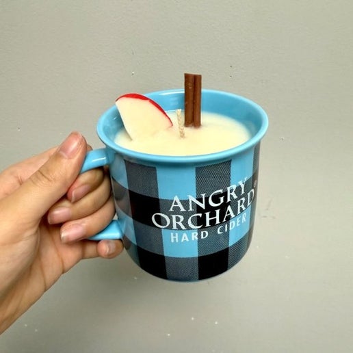 Hand holding a blue and black plaid mug with a candle in it. On the front of the mug in white text it reads "Angry Orchard Hard Cider". The candle scent is Hot Apple Cider, containing a faux cinnamon stick and apple slice for decoration.