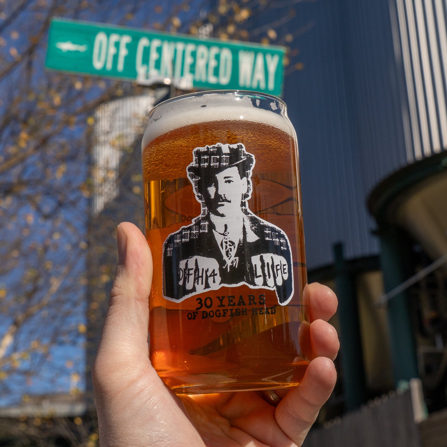 A hand holds an Off Centered Way Can Glass by Grandstand Glassware & Apparel, filled with amber beer and featuring a “30 Years” logo, in front of a green “Off Centered Way” sign and a modern building under a blue sky.
