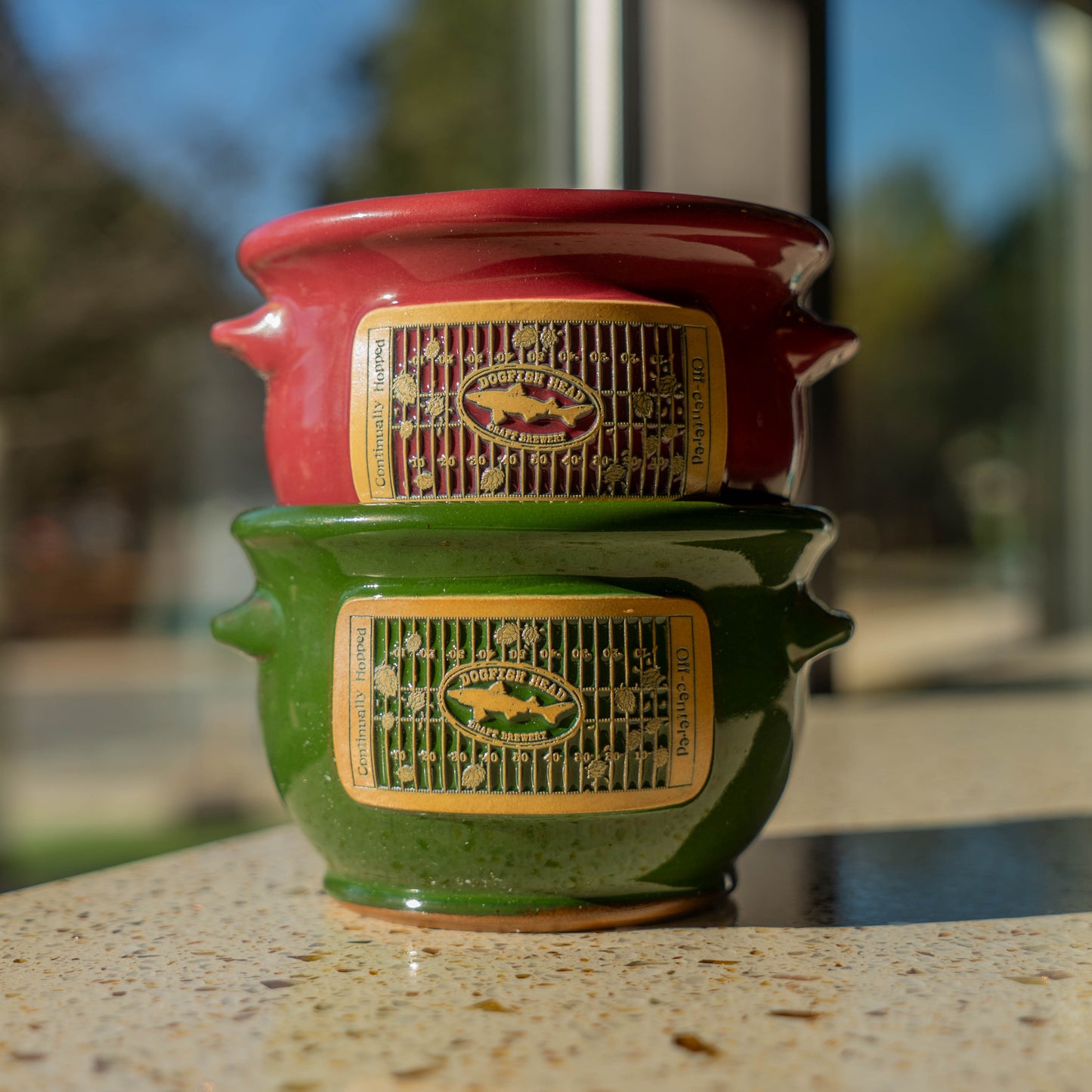 Stack of red and green ceramic crocks with branding on a blurred outdoor background
