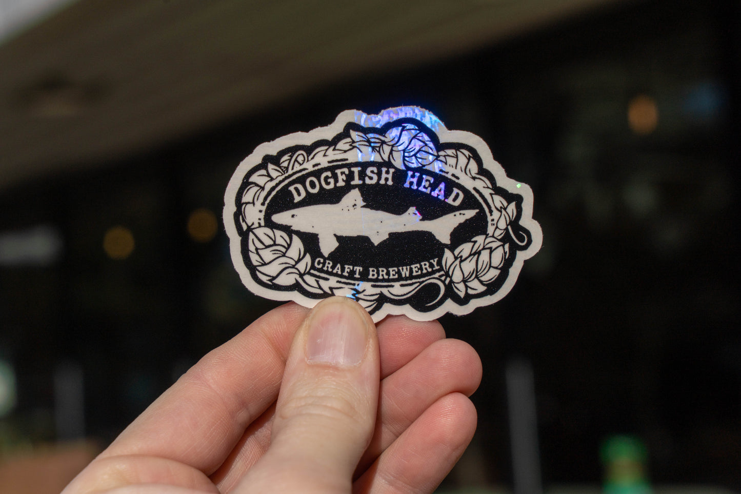 A hand holds the Suncatcher Logo Sticker by Rainbow Symphony, featuring a holographic design of a shark and hop vines—part of the Dogfish Head Craft Brewery logo—on a black and white background.