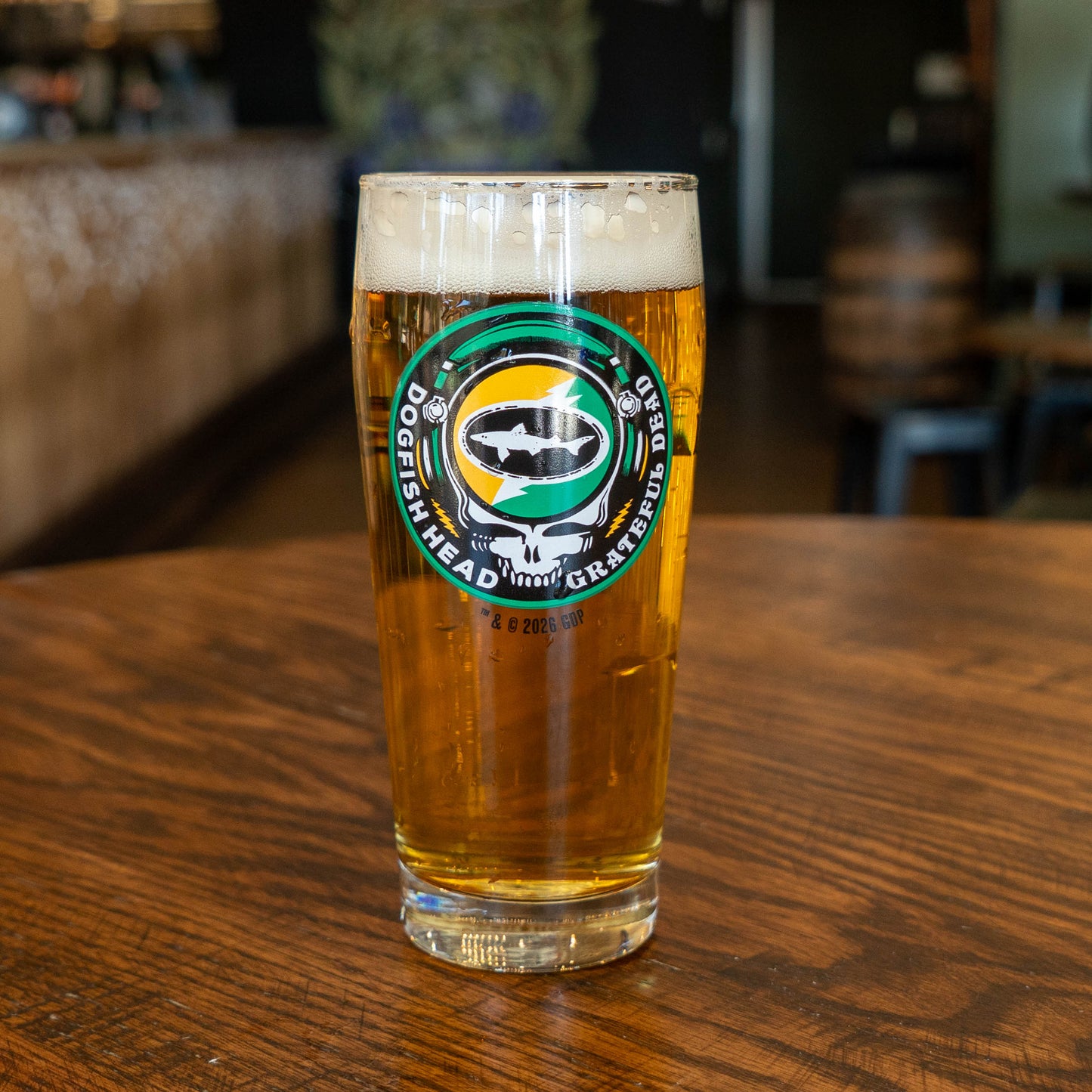 A pint of amber beer in a Dogfish Head x Grateful Dead Citrus Daydream Glass by Grandstand Glassware & Apparel rests on a wooden table in a bar or brewery setting.