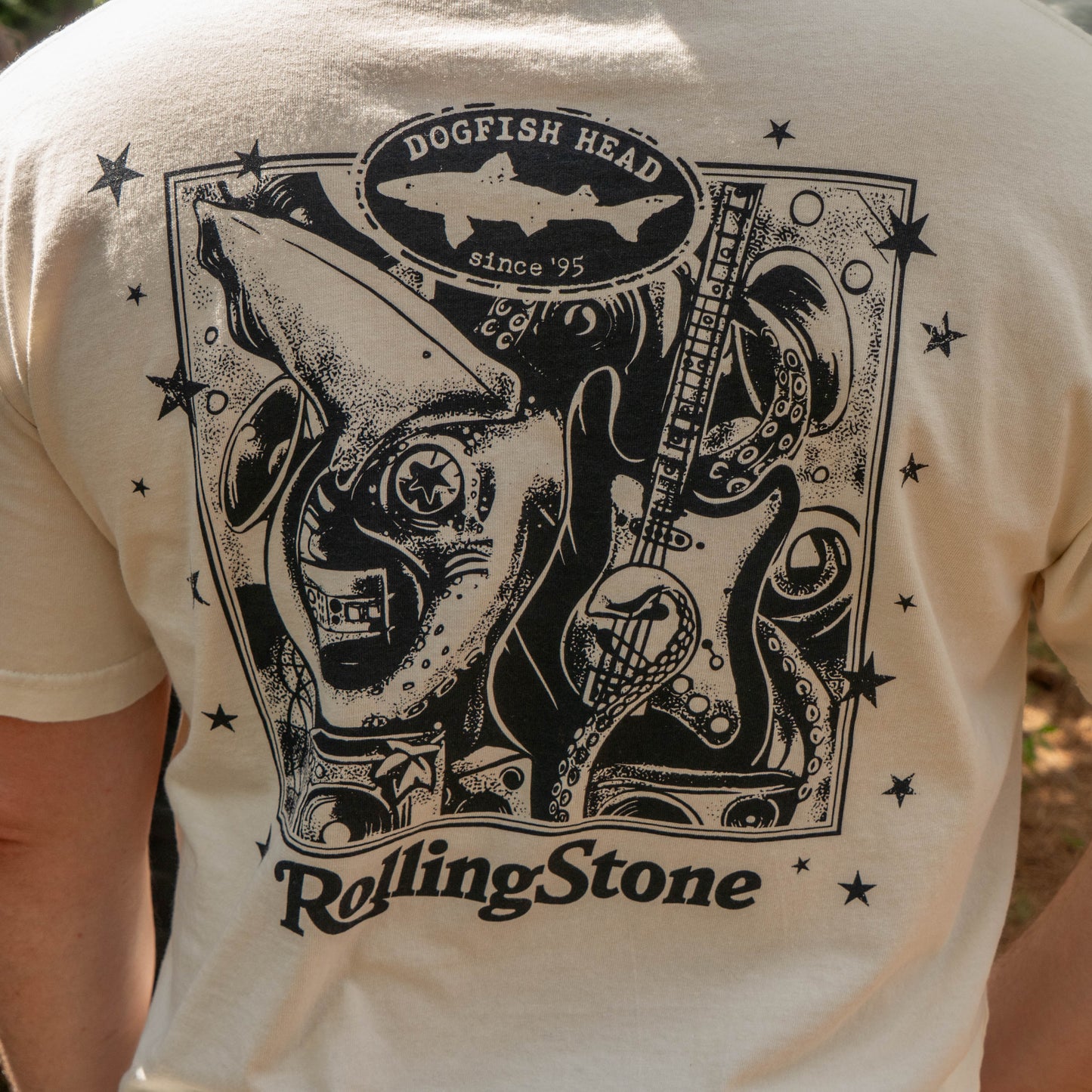 The back of the cream-colored Dogfish Head x Rolling Stone Oatmeal Tee Shirt features a black graphic with a shark, guitar, stars, and "Dogfish Head since 95" and "Rolling Stone.