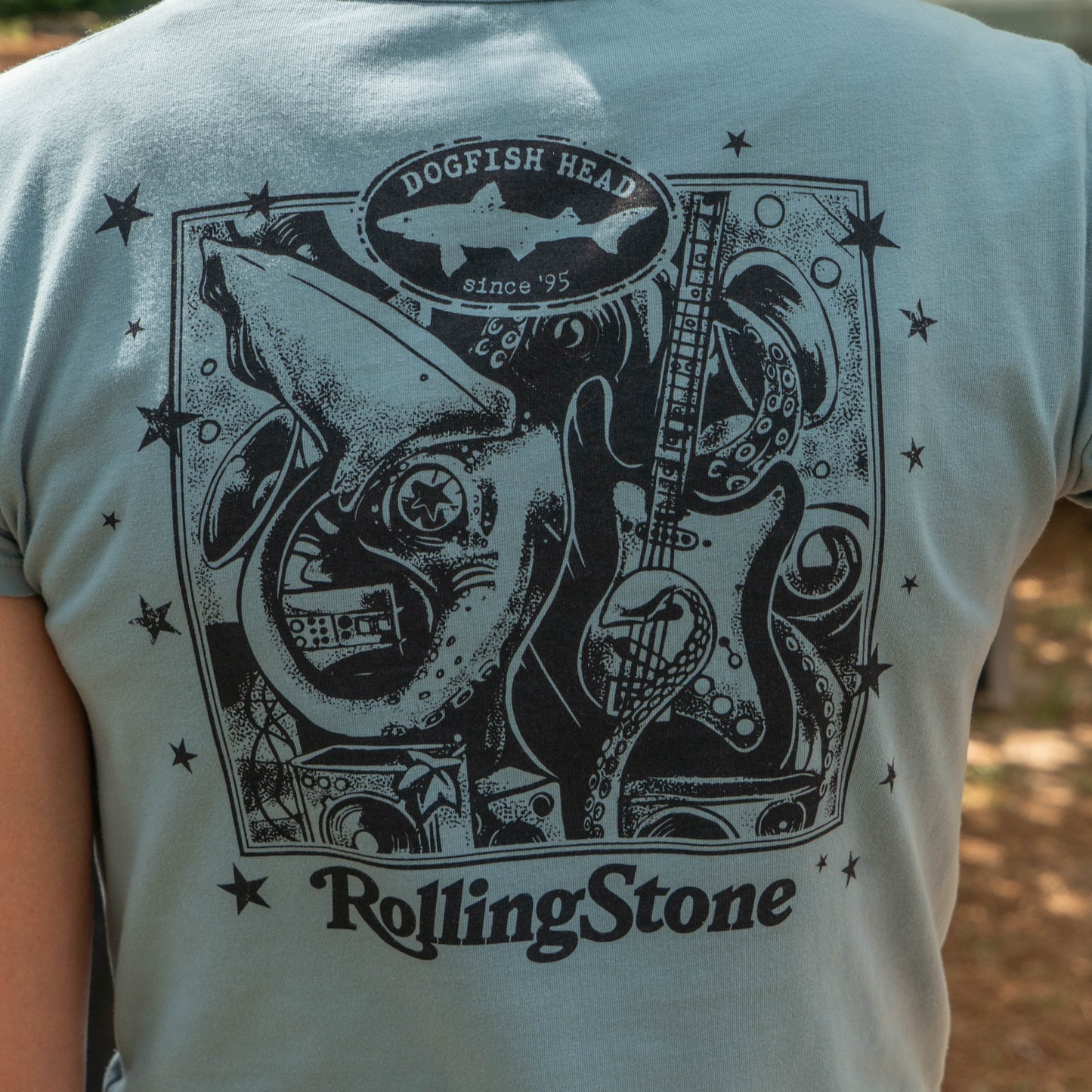 A woman wears the Dogfish Head x Rolling Stone Women's Slate Blue Baby Tee Shirt by GO WEST T-SHIRT COMPANY, a slim fit tee featuring an artistic squid, guitar, stars, and "Dogfish Head since 95" with "Rolling Stone" text on the back.
