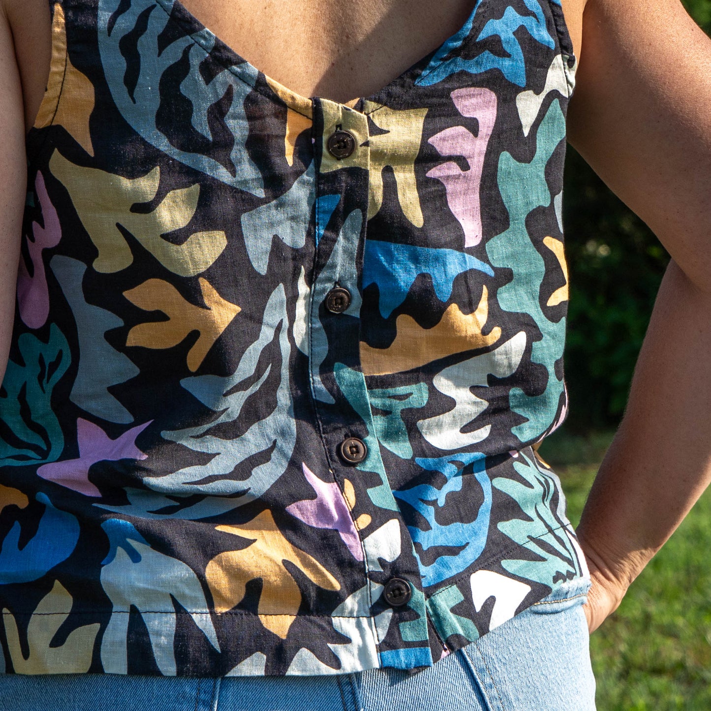 Wearing the Patagonia W Garden Island Tank Top, a vibrant abstract-patterned reversible top with back buttons by Patagonia, paired with light blue jeans, the person stands outdoors hands on hips.
