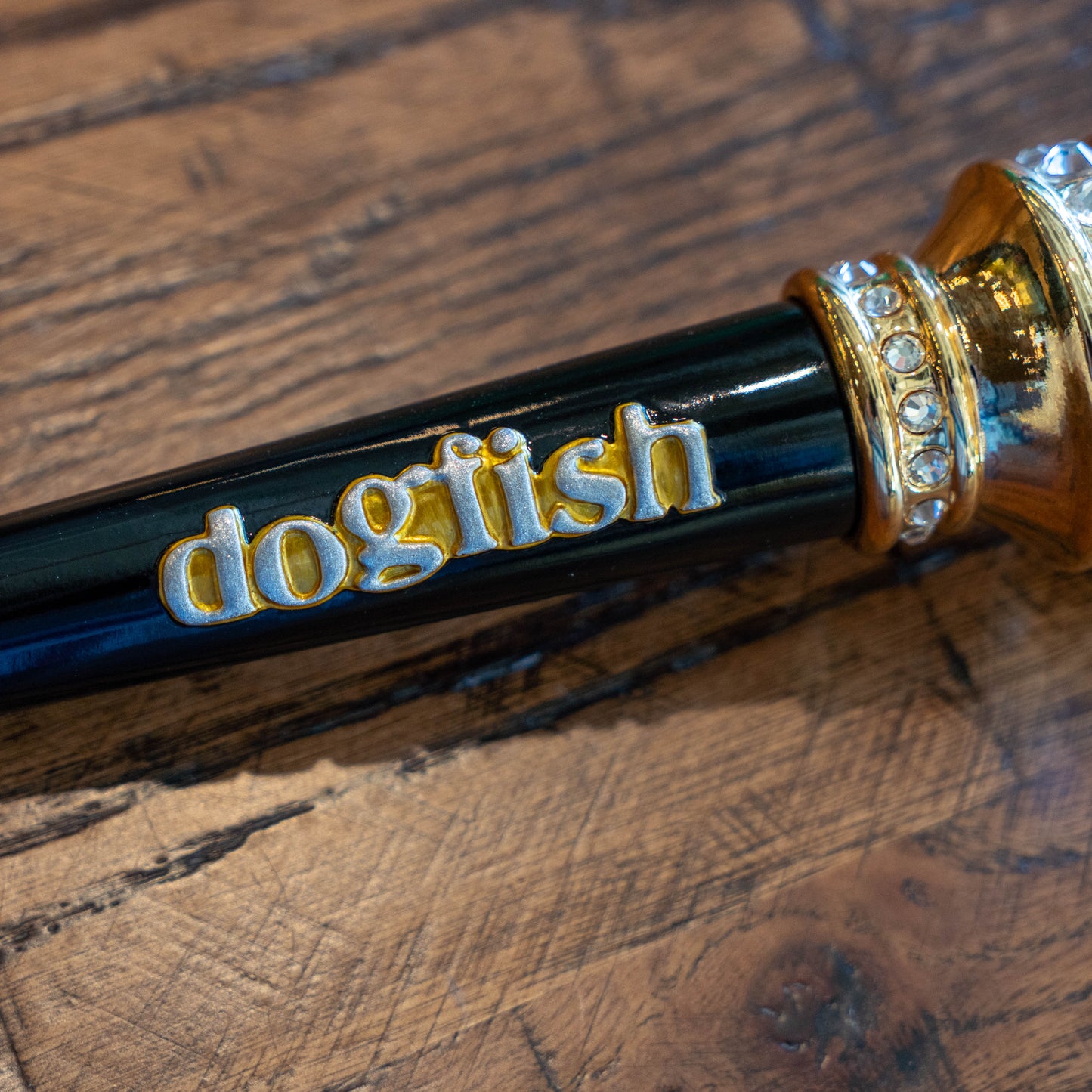 A close-up of a Taphandles Inc. OG Uber Tap Handle in black and gold, featuring "dogfish" in yellow and white lettering, resting on a textured wooden surface.
