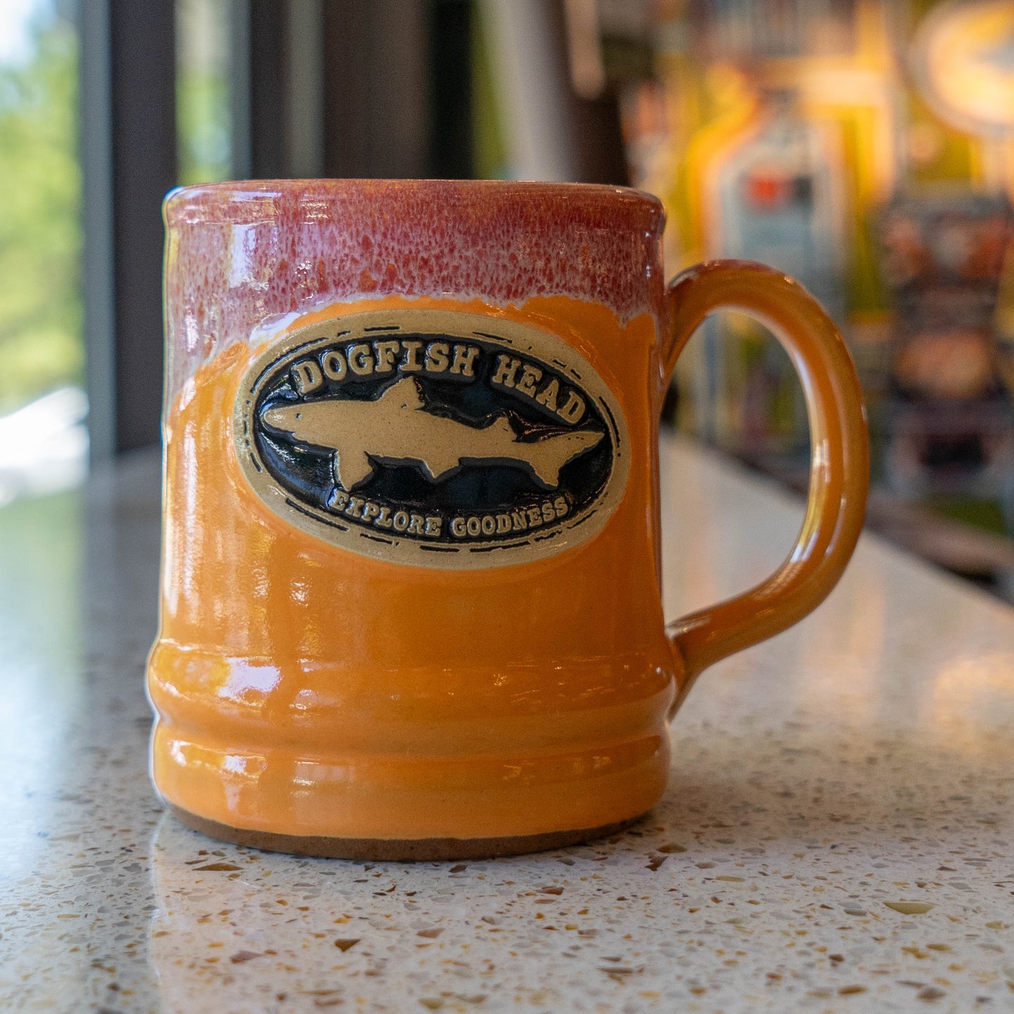 A Tangerine Ramsey Core 14oz ceramic mug by Deneen Pottery with a handle sits on a speckled countertop. It features an oval logo with two fish silhouettes and “DOGFISH HEAD EXPLORE GOODNESS” on the front.