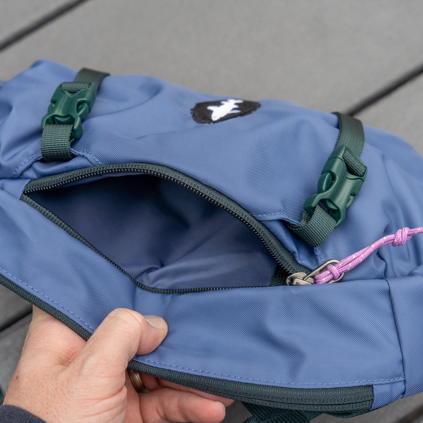 A hand opens the zippered pocket of a Patagonia Blue Atom Sling 8L, featuring green buckles and a pink cord. The empty, recycled polyester interior is visible as the bag rests on a gray wooden surface.