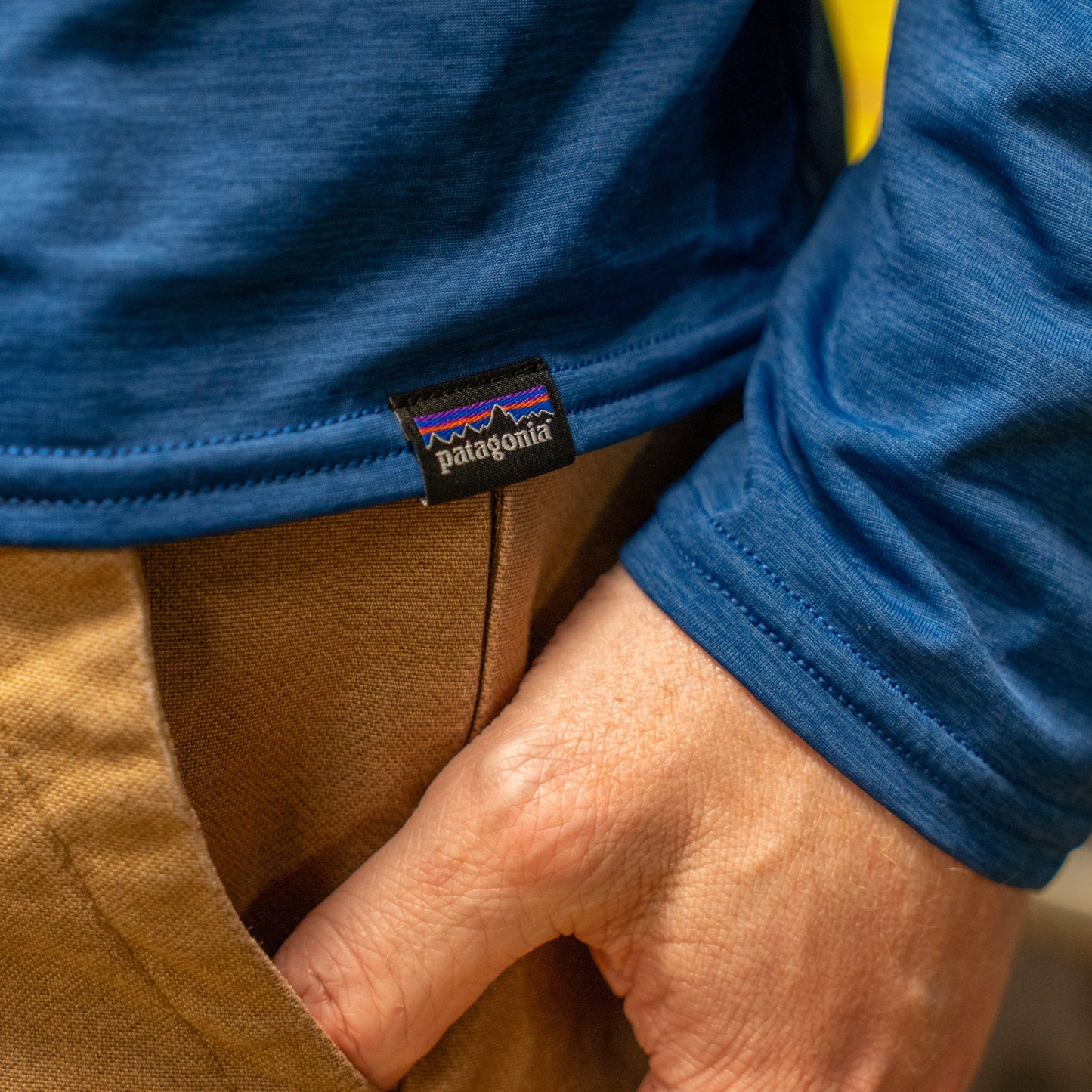 A person wears a Patagonia M Blue Long Sleeve Runner shirt, made from recycled polyester, with brown pants. Their right hand is partly in their pocket, and the visible Patagonia logo tag is on the shirt hem.