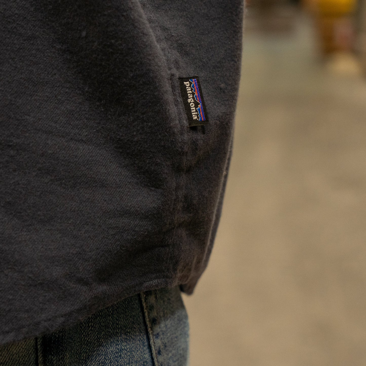 Close-up of a person wearing a Patagonia M Ink Blue Light Weight Flannel Button Up and blue jeans, highlighting the small black Patagonia tag on the shirt’s side seam. The background is blurred.