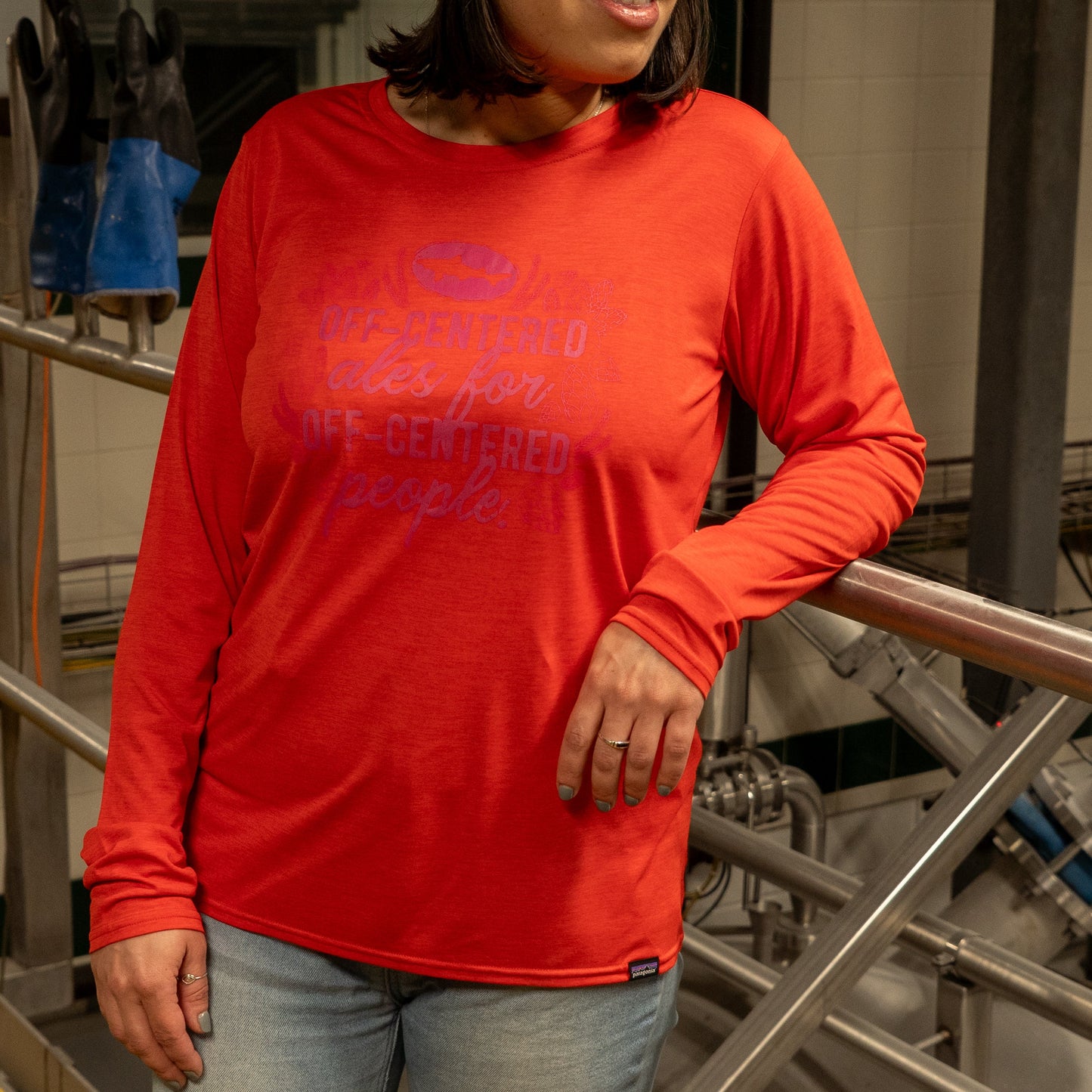 A person in a bright red Patagonia W Red Long Sleeve Runner shirt stands indoors, one arm on a metal railing. Industrial equipment and gloves hang in the background, with the person's face partially out of frame.