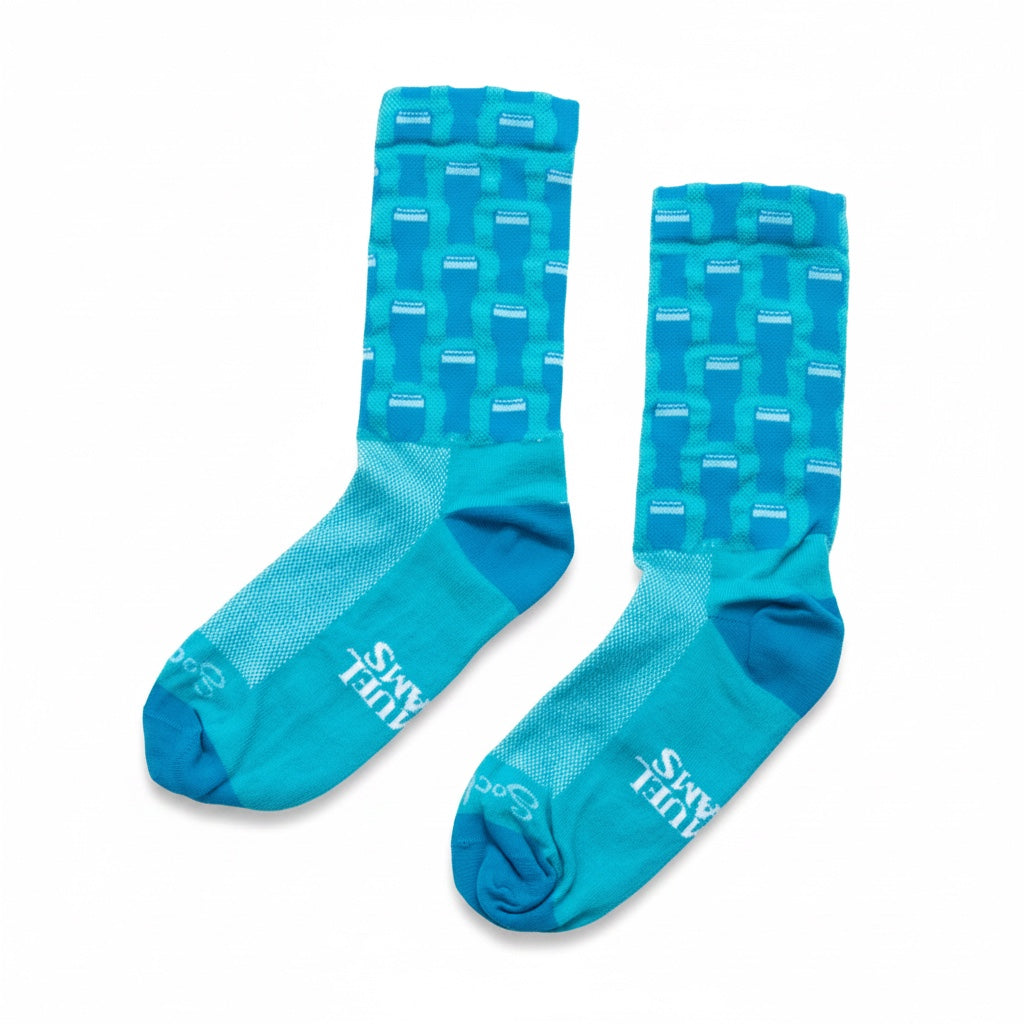 A pair of athletic crew socks in bright turquoise blue with a lighter blue geometric pint pattern on the upper leg and white 'Sock Guy' branding on the toes, white 'Samuel Adams branding on the foot, shown against a plain white background.