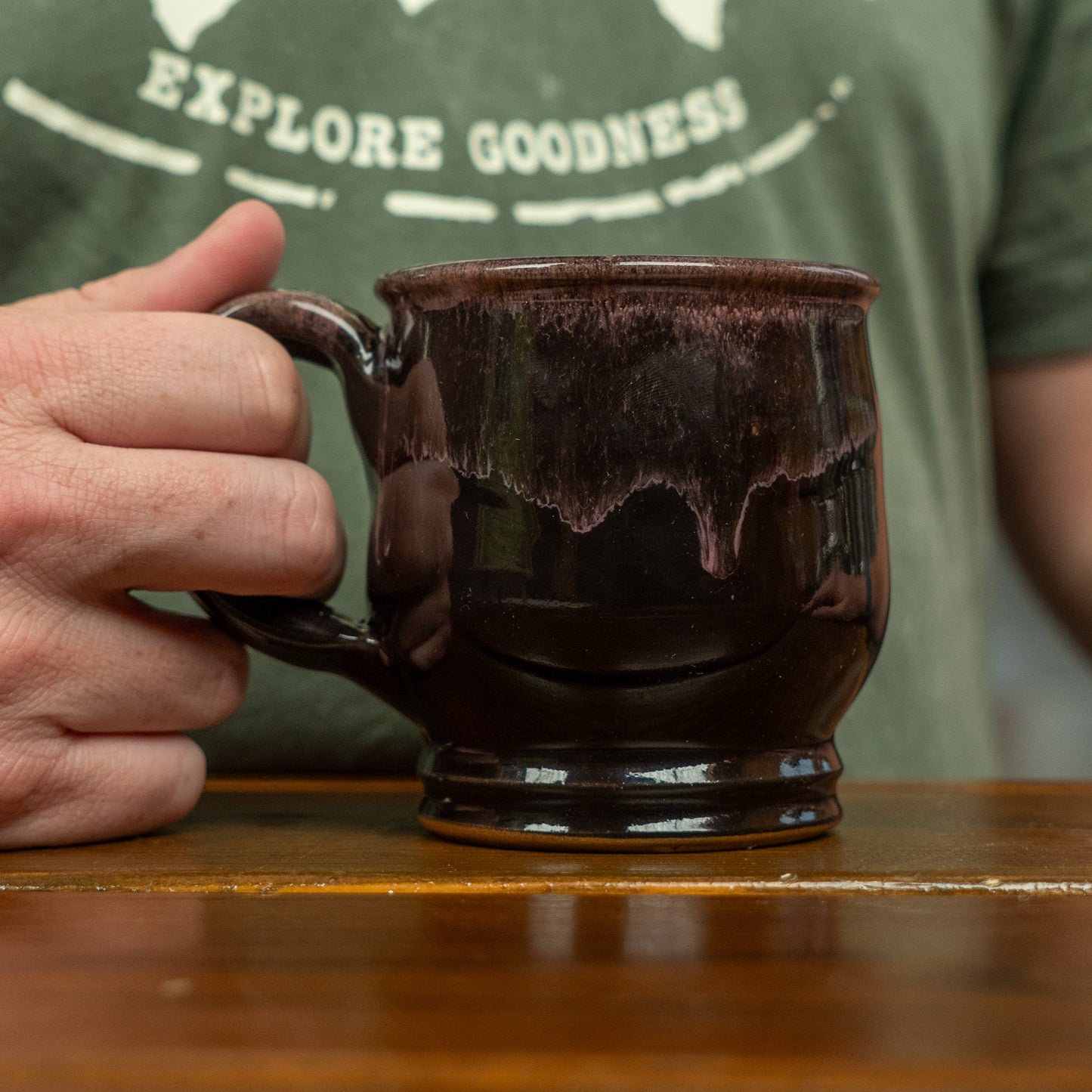 A person in a green shirt with "EXPLORE GOODNESS" text holds a Black Punkin Mug by Deneen Pottery on a wooden surface, highlighting their unique seasonal drinkware.