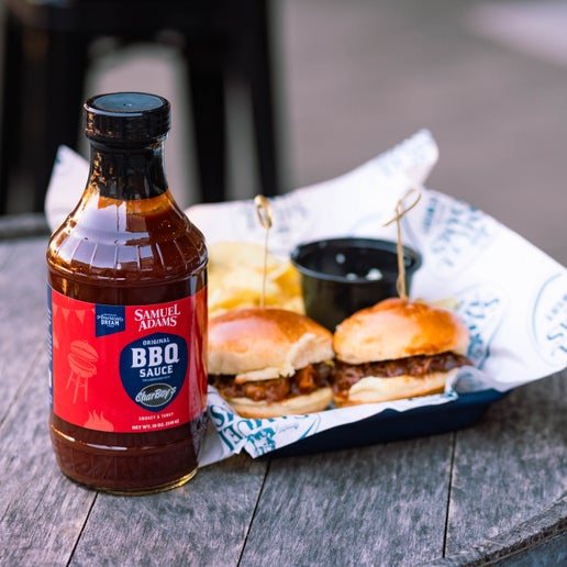 The Sam Adams x CharBoy's BBQ Sauce bottle shown in front of two sandwiches