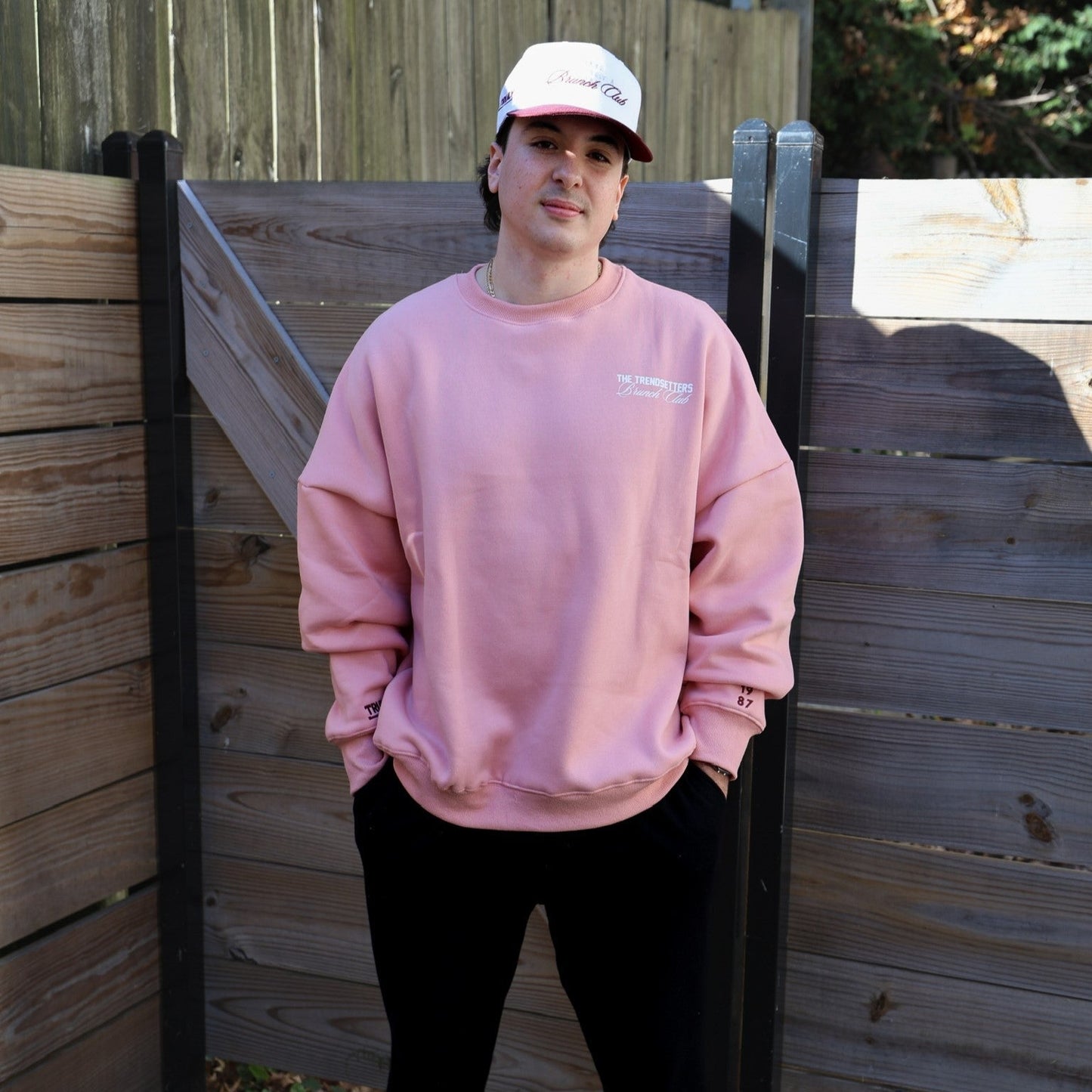 Front of a person standing outdoors, wearing an oversized pink crewneck sweatshirt, black pants, and a white and maroon baseball cap. The sweatshirt has 'THE TRENDSETTER Brunch Club' in white text on the left chest, and the numbers '19' and '87' in maroon on the right cuff. The baseball cap has a pink script logo that says 'Brunch Club'. The person has their hands in their pockets and is smiling slightly, standing in front of a rustic wooden fence.