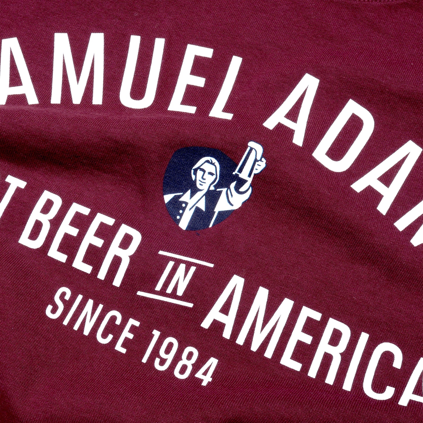 Close-up of graphics on the front. Front view of tee features writing "Samuel Adams" a small image of blue shield with Sam Adams hoisting a beer and writing "Best Beer in America Since 1984" underneath.