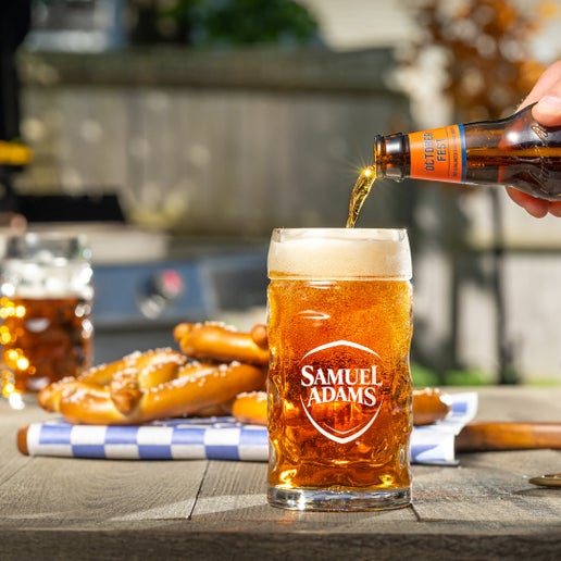 A half liter Octoberfest Glass Stein with a white Sam Adams logo on it, modeled after authentic Bavarian beer glassware sitting on a wooden table with a beer being poured into it in front of a traditional Octoberfest background. Glass is made of dimpled heavy duty glass.