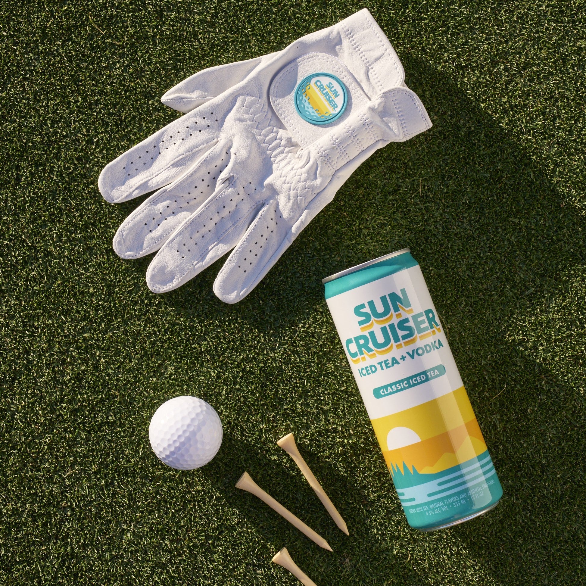 Sun Cruiser x Gfore golf glove, Sun Cruiser classic iced tea, and a golfball and tees on a grass background
