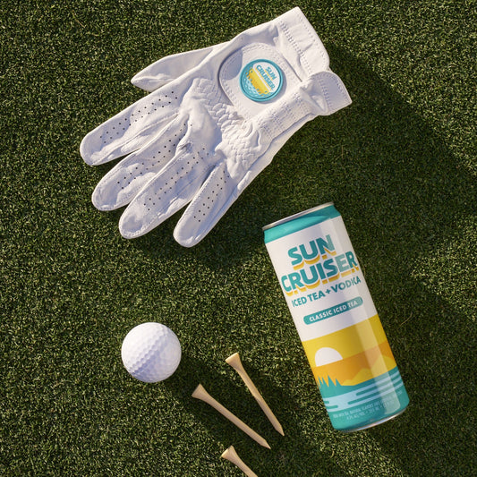 Sun Cruiser x Gfore golf glove, Sun Cruiser classic iced tea, and a golfball and tees on a grass background