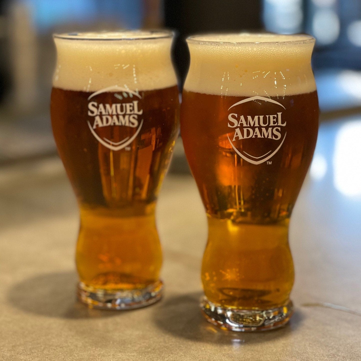 Two 16oz pint glasses filled with beer with the Samuel Adams classic shield logo printed on the front in white.