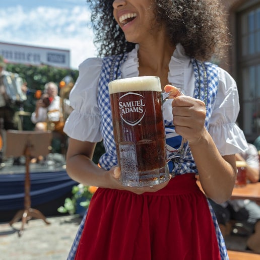 A woman in a dirndl holding a beer-filled 1L Glass Stein traditional for Octoberfest, with a glass handle and the classic Samuel Adams logo printed on the front in white.