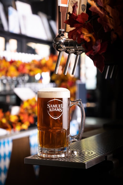 A beer-filled 1L Glass Stein traditional for Octoberfest, with a glass handle and the classic Samuel Adams logo printed on the front in white, sitting on a bar under a beer tap.