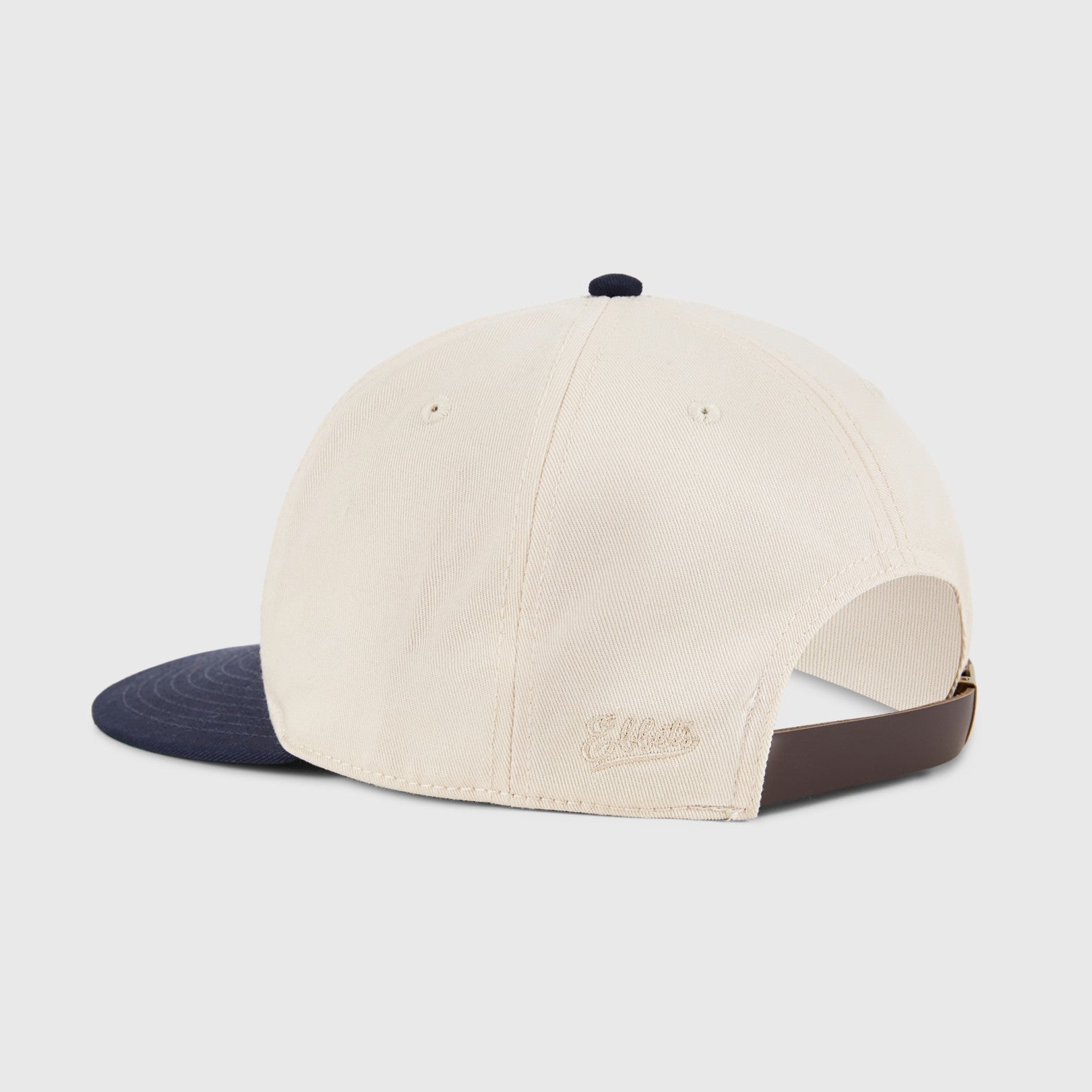 Beige cap with navy brim and brown strap on a light gray background