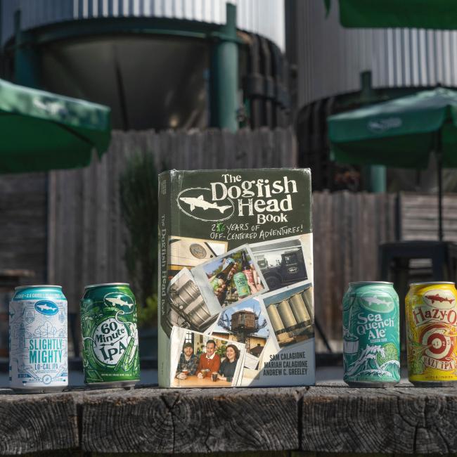 Four vibrant Dogfish Head beer cans and the "Dogfish Head Book: 26 Years of Off-Centered Adventures" by J Wiley and Sons Publishing are displayed outdoors, with green umbrellas and tanks in the background—a treat for craft beer fans.
