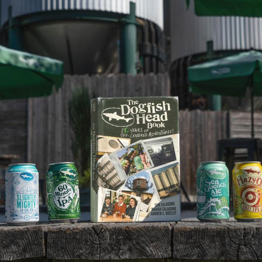Four vibrant Dogfish Head beer cans and the "Dogfish Head Book: 26 Years of Off-Centered Adventures" by J Wiley and Sons Publishing are displayed outdoors, with green umbrellas and tanks in the background—a treat for craft beer fans.