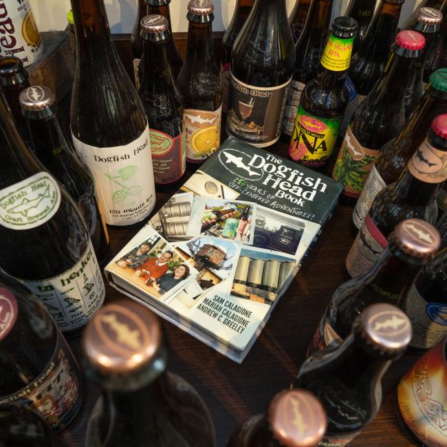 Several Dogfish Head Craft Brewery beers are arranged around the "Dogfish Head Book 26 Years of off Centered Adventures" by J Wiley and Sons Publishing, capturing the innovative spirit of craft beer.