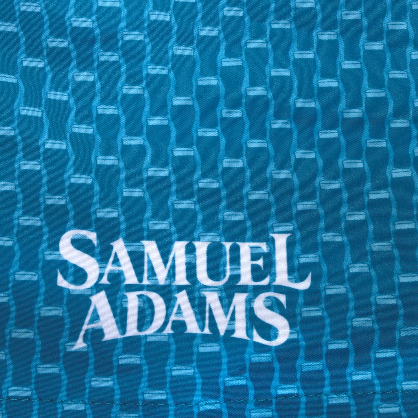 Close-up of Samuel Adams swim trunks featuring blue pint glass pattern fabric and white logo print on quick-dry material