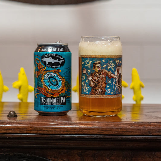 A Grandstand Glassware & Apparel 75 Minute Can Glass filled with frothy beer sits on a wooden surface, with four yellow banana-shaped figurines in the background.