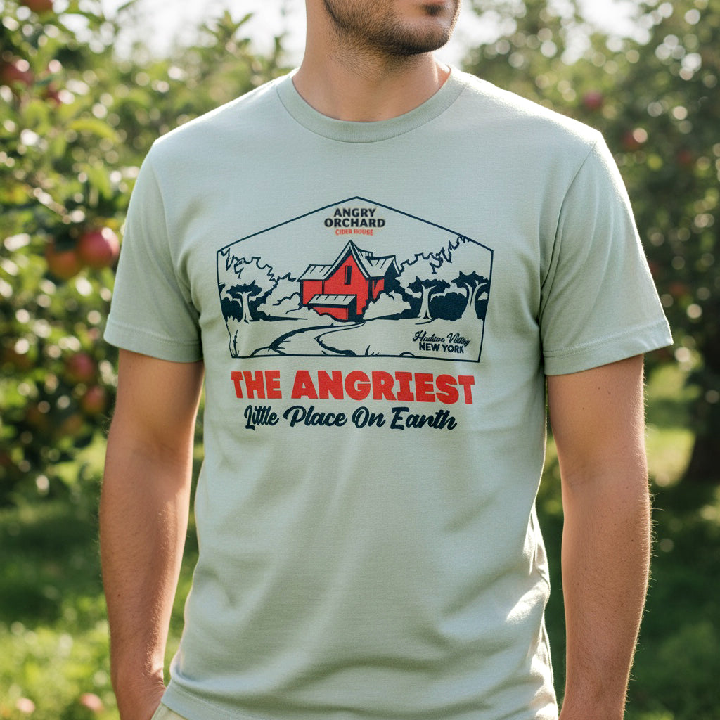 A man wearing a sage green graphic t-shirt in an outdoor apple orchard setting. The shirt features a red and navy blue graphic of a barn with the text 'ANGRY ORCHARD' and 'THE ANGRIEST Little Place On Earth'.