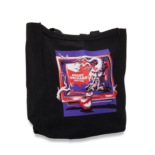 A black canvas tote bag featuring a colorful graphic print of an angry tree emerging from a red tv with the text 'ANGRY ORCHARD' on a white background.