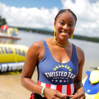 A smiling woman with braided hair stands on a sandy shore, wearing a one-piece swimsuit with a dark blue top featuring the "TWISTED TEA HARD ICED TEA" logo, a yellow sunburst, and five white stars. The bottom of her suit has red and white stripes. Behind her, a yellow inflatable "TWISTED TEA KEEP IT TWISTED" buoy floats in the water to the left, and a blue and yellow jet ski is partially visible on the right.