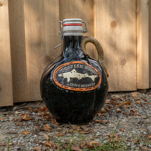 A round, dark brown ceramic Blk Punkin Growler by Romanick Pottery with a handle and flip-top lid sits on gravel near a wooden fence, surrounded by dappled sunlight and fallen leaves.