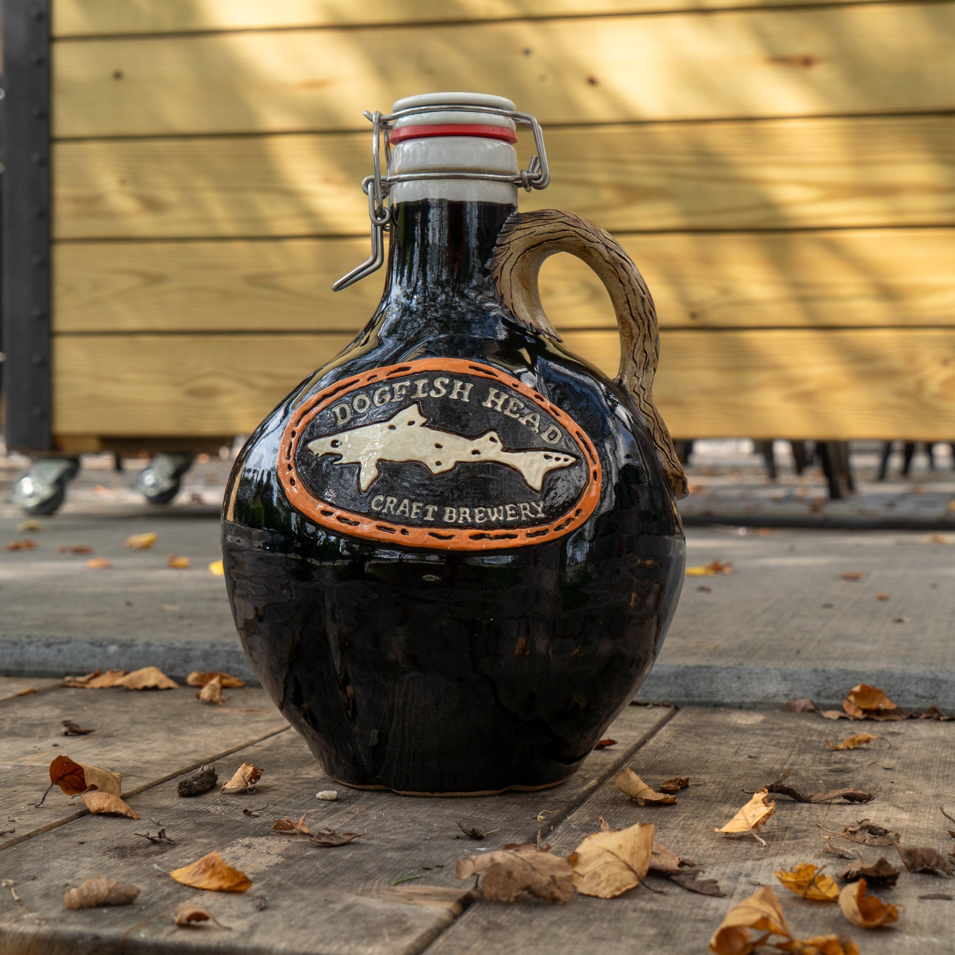 A dark glass Blk Punkin Growler by Romanick Pottery, with a handle and swing-top lid, sits on a wooden surface outdoors amid dry autumn leaves. A wooden fence stands in the background.