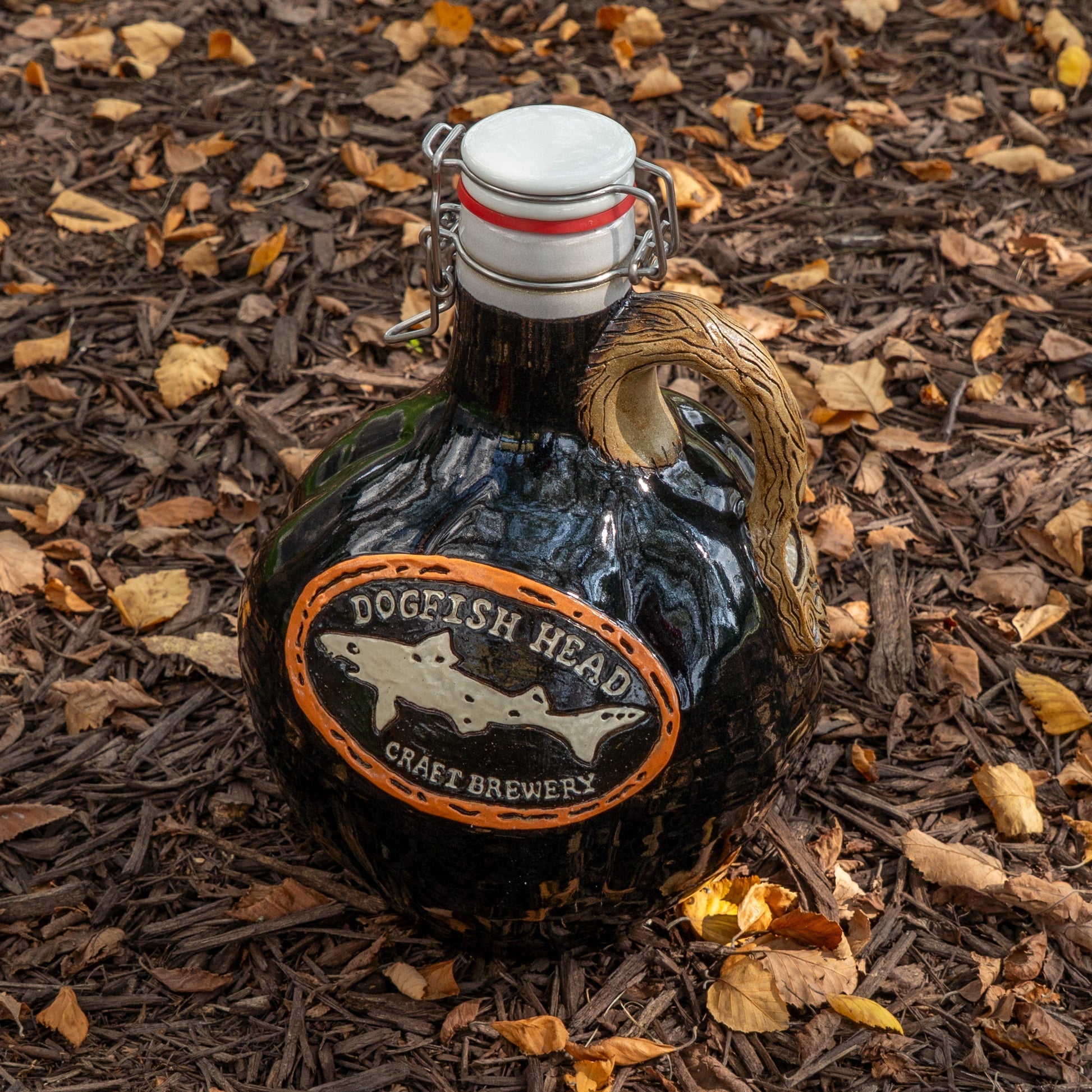 A brown ceramic Blk Punkin Growler by Romanick Pottery, featuring a handle and metal clasp lid, rests on mulch with autumn leaves—ideal for handmade growler enthusiasts and fans of seasonal brews alike.