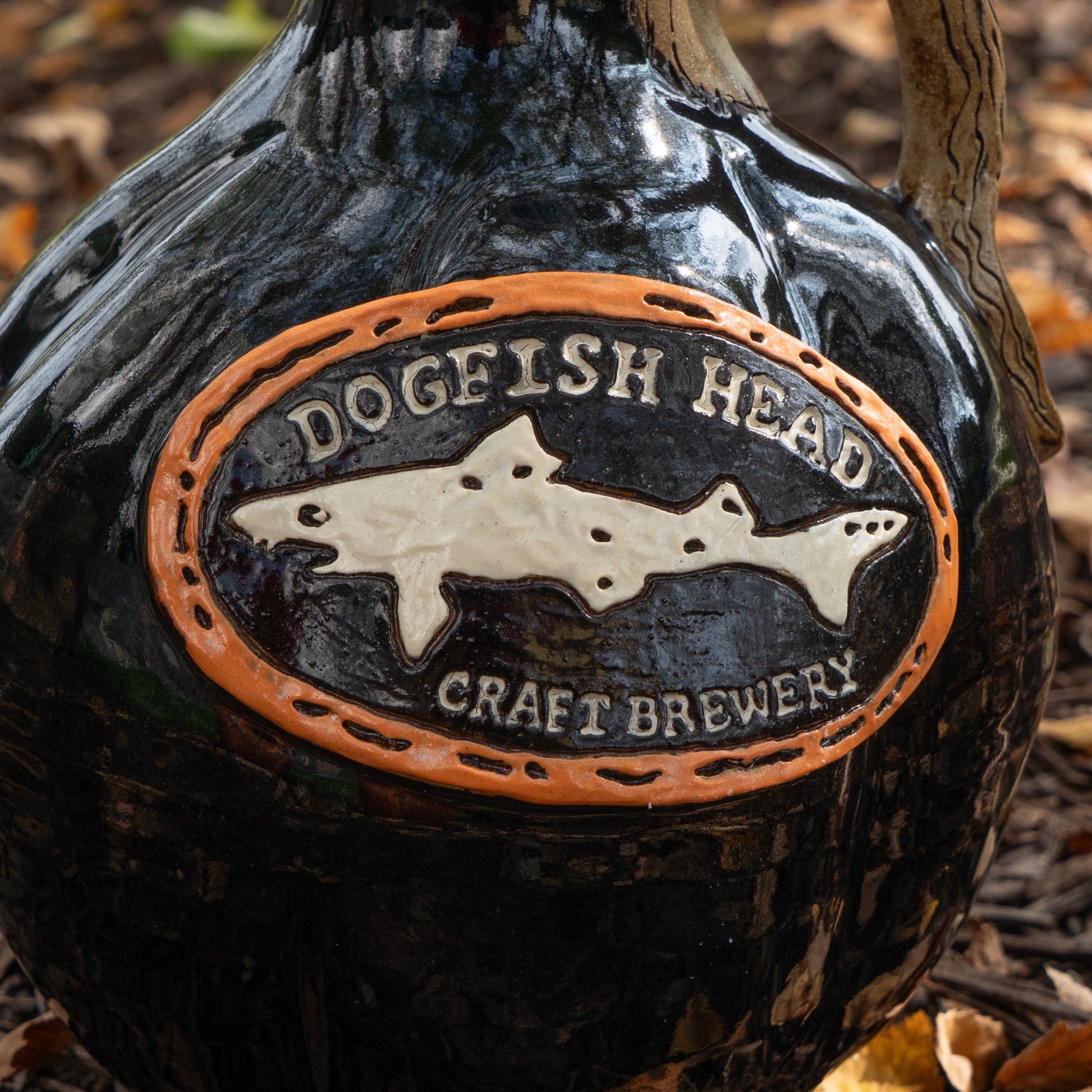 A close-up of the Blk Punkin Growler by Romanick Pottery displays its embossed logo—a dogfish in an orange oval—set against a glossy black finish with leaves and mulch, highlighting the artisanal craftsmanship.