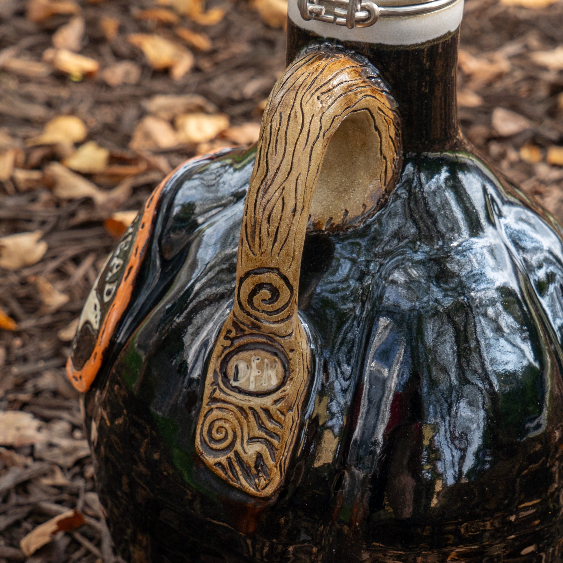 A close-up of the Blk Punkin Growler by Romanick Pottery—a dark, glossy ceramic jug with a textured, spiral-carved handle and "OPEN" inscribed—rests on bark mulch with dry leaves in the background.
