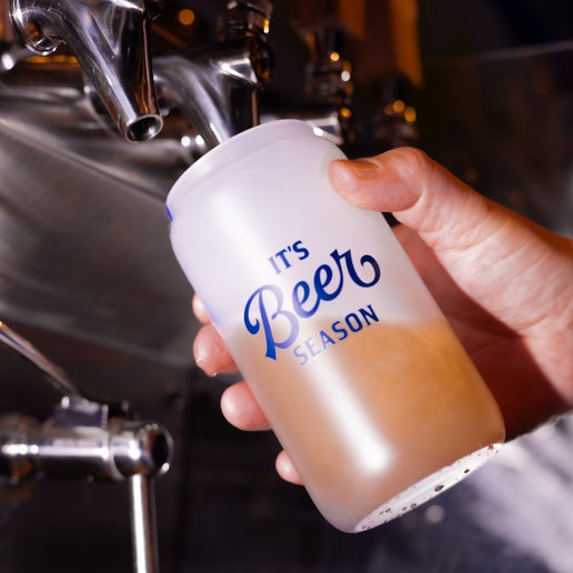A frosted glass with the phrase "It's Beer Season" in navy printed on one side of it that changes color to a blue when it comes into contact with cold. In this image the glass is being filled with beer from a tap.