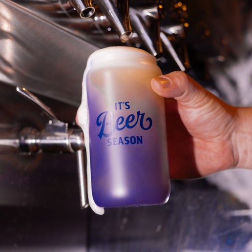 A frosted glass with the phrase "It's Beer Season" in navy printed on one side of it that changes color to a blue when it comes into contact with cold. In this image the glass has just been filled with beer from a tap and the glass is actively changing color to blue.