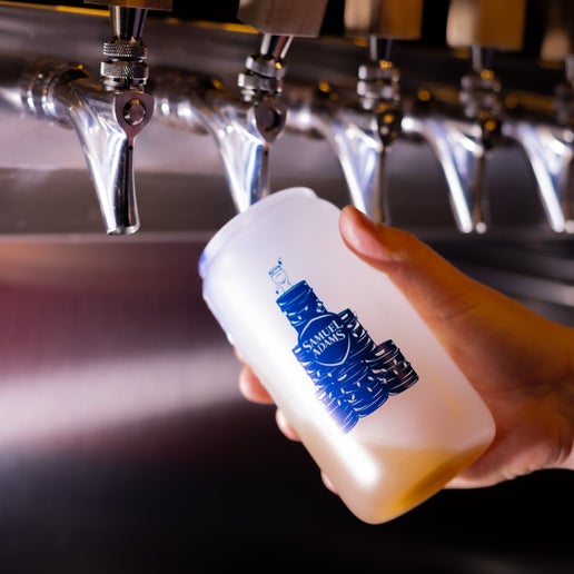 A frosted glass with a pyramid of kegs wrapped in Christmas lights with a perfect pint glass sitting on top and the Samuel Adams classic shield logo in the middle in navy printed on the other side of it that changes color to a blue when it comes into contact with cold. In this image the glass is being filled up with beer from a tap.